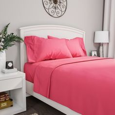 a bed with pink sheets and pillows next to a white night stand in front of a window