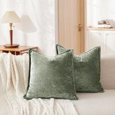 two green velvet pillows sitting on top of a white couch next to a wooden table