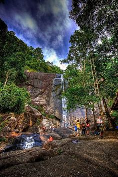 people standing in front of a waterfall surrounded by green trees and rocks, under a cloudy blue sky