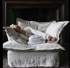 a woman laying on top of a white couch covered in pillows and blankets with her head resting on the pillow