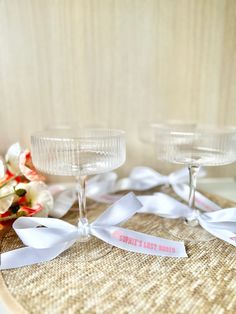 two wine glasses sitting on top of a table with white ribbon around them and flowers in the background