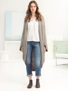 a woman standing in front of a white wall wearing jeans and a cardigan sweater