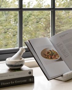 an open book sitting on top of a table next to a bowl of food near a window