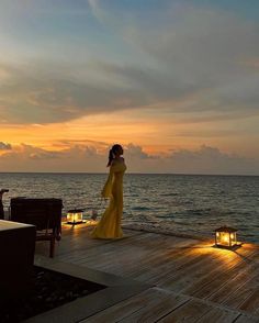 a woman in a long yellow dress standing on a dock at sunset with candles lit