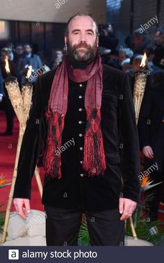 a bearded man in a black suit and red scarf standing on a red carpet with torches