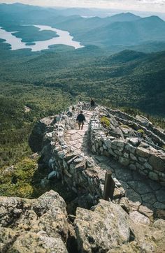 two people walking up the side of a stone wall on top of a mountain with mountains in the background