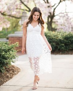 a woman wearing a white dress and heels walking down a sidewalk with trees in the background