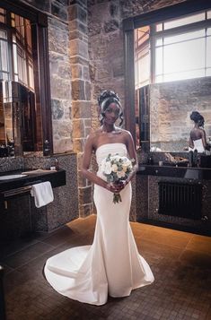 a woman in a white wedding dress is standing near a mirror and holding a bouquet