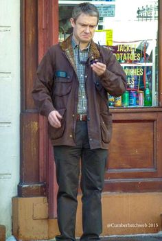 a man standing in front of a store looking at his cell phone while wearing a brown jacket