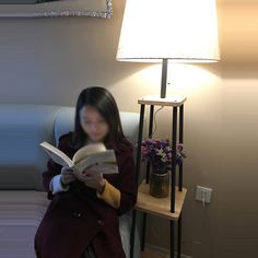 a woman sitting in a chair reading a book next to a lamp and flower pot