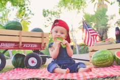 a baby sitting on the ground eating watermelon next to a wagon and american flags