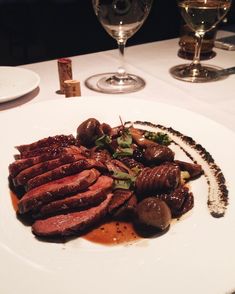 a white plate topped with meat and veggies on top of a table next to wine glasses