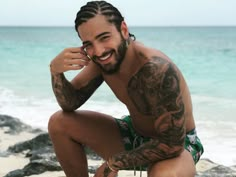 a man with tattoos on his arm and chest squatting in front of the ocean