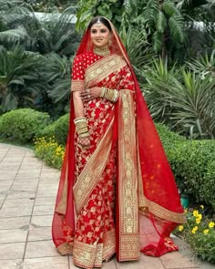 a woman in a red and gold bridal gown standing on a brick walkway with greenery behind her