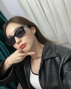 a woman in black leather jacket and sunglasses holding her hand near her face with both hands