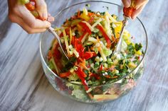 two hands are holding tongs over a salad in a glass bowl on a wooden table