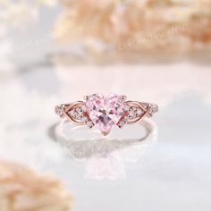 a pink heart shaped diamond ring on top of a white surface with flowers in the background