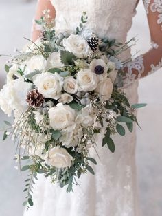 a bride holding a bouquet of white roses and greenery in her hands, with pine cones on the side