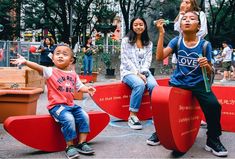 three children sitting on red benches in the middle of a city park with people standing around