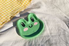 a green frog shaped bowl sitting on top of a white bed sheet next to a yellow checkered pillow