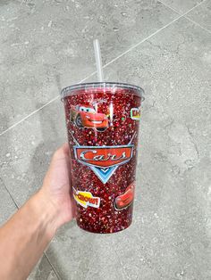 a hand holding a cup with cars on it and the lid is covered in red glitter