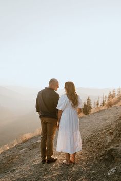 a man and woman standing on top of a hill looking at the sky with mountains in the background