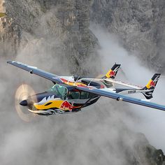 an airplane flying in the air with mountains behind it and fog rolling around below,