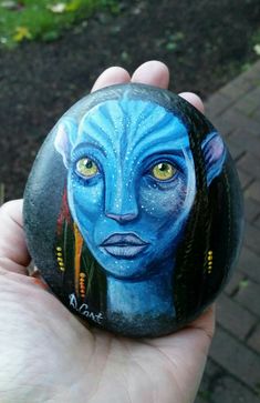 a hand holding a painted rock with an avatar on it's face and yellow eyes