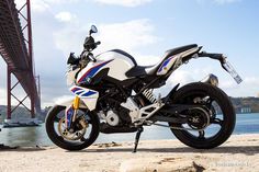 a white and blue motorcycle is parked on the sand by the water with a bridge in the background