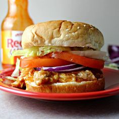 a chicken sandwich with lettuce, tomato and onion on a red plate next to a beer bottle