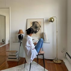 a woman sitting on a chair in front of a wall with a painting and lamp