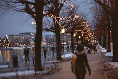 people are walking down the sidewalk near trees with lights on them and snow covered ground