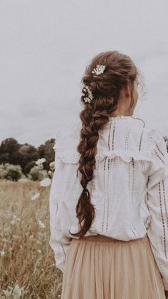 a girl with long hair standing in a field wearing a white blouse and tan skirt