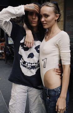 a man and woman standing next to each other in front of a building with a skull on it's shirt