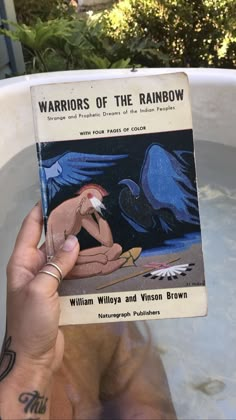 a person holding up a book in front of a tub full of water and plants