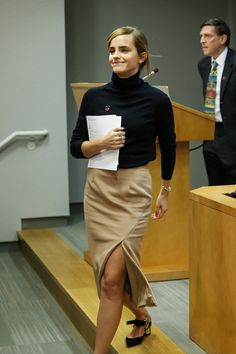 a woman in a black shirt and tan skirt is standing at a podium while holding a piece of paper