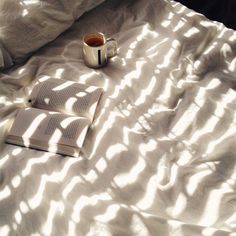 an open book and cup of coffee sit on a bed with white sheets in the sunlight