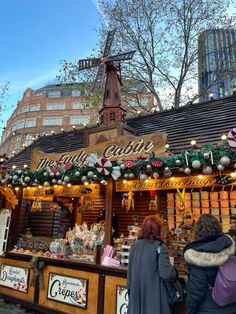 two women are standing in front of a christmas market stall with lights and decorations on the roof