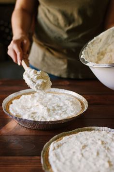 two pie pans filled with white frosting on top of a wooden table next to a person holding a spatula