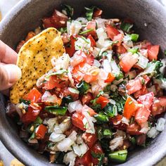 a hand holding a tortilla chip over a bowl filled with salsa and vegetables