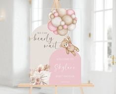 a welcome sign with a teddy bear and balloons hanging from it's side on an easel