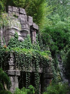 a stone statue with vines growing on it's face next to a small waterfall