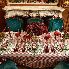 the table is set for christmas dinner with red flowers and candles on it, along with green velvet chairs