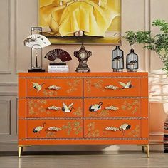 an orange dresser with birds on it in front of a painting and fan next to potted plant