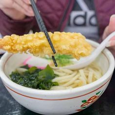 a person holding chopsticks over a bowl of noodles with greens and sauce on the side