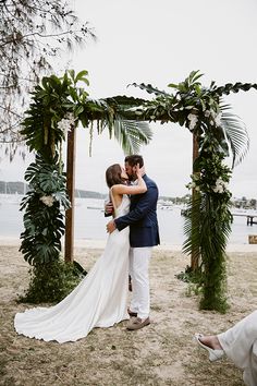 a man and woman kissing under an arch with greenery on the beach in front of them