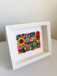 a white frame with colorful paper flowers in it on a table next to a wall