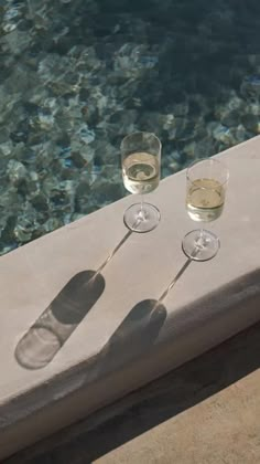 two glasses of wine are sitting on a ledge near the water's edge, next to a swimming pool