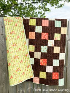 two quilts hanging on a wooden fence in front of a green leafy tree