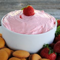 a white bowl filled with pink frosting surrounded by cookies and strawberries